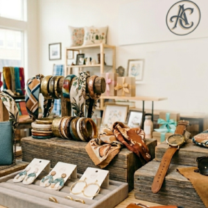 A stylish boutique display featuring fashion accessories including handbags, bracelets, scarves, earrings, watches, sunglasses, and jewelry arranged on rustic wooden stands inside a modern lifestyle store with bright natural lighting.