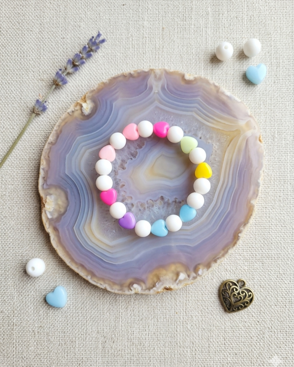 Cute colorful heart bead bracelet with pastel pink, blue, yellow, green, and purple heart-shaped beads displayed inside a transparent jewelry display box on a wooden surface with decorative flowers and accessories.