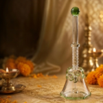 Clear crystal glass Om pooja bell with a green finial set on a golden cloth beside a glowing brass diya lamp and marigold flowers.
